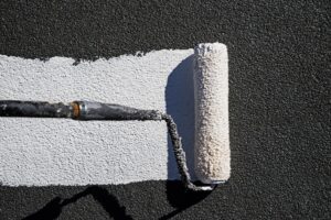 Acrylic Roof Coating 1 A paint roller applies a strip of white elastomeric roof coating to an aggregate roof