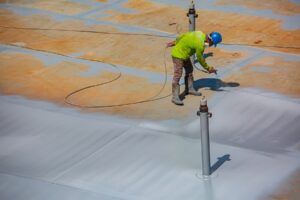 A worker in safety gear sprays silicone roof coating onto a large flat roof with pipes