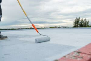 A person uses a paint roller with a long handle to apply acrylic roof coating to a flat rooftop under a cloudy sky.