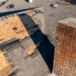 A roof with storm roof damage—missing and damaged shingles, exposed plywood, and a large brick chimney in the foreground.